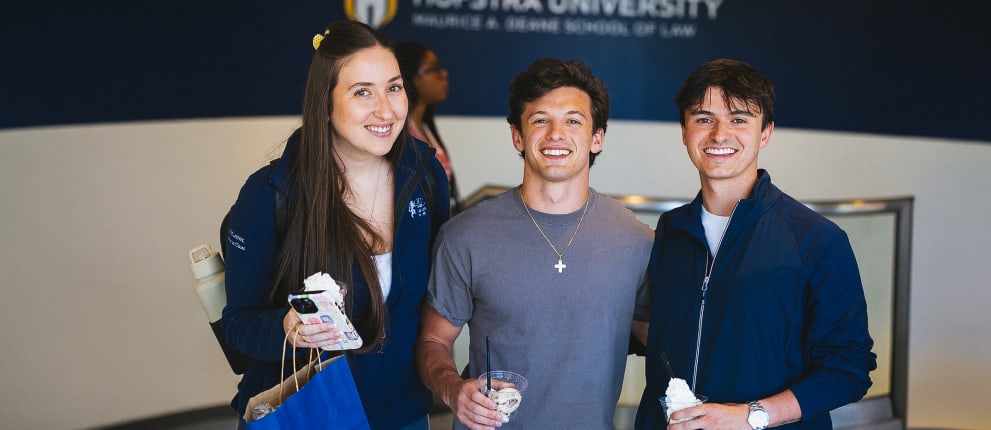Three young adults smiling and holding ice cream cups inside a building with a Hofstra University School of Law sign in the background. One holds a blue gift bag and a drink bottle.