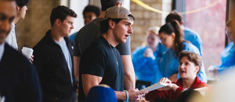A group of people gather indoors, some standing and talking, while others sit and engage in discussion. The setting appears casual, with a mix of relaxed and focused expressions.