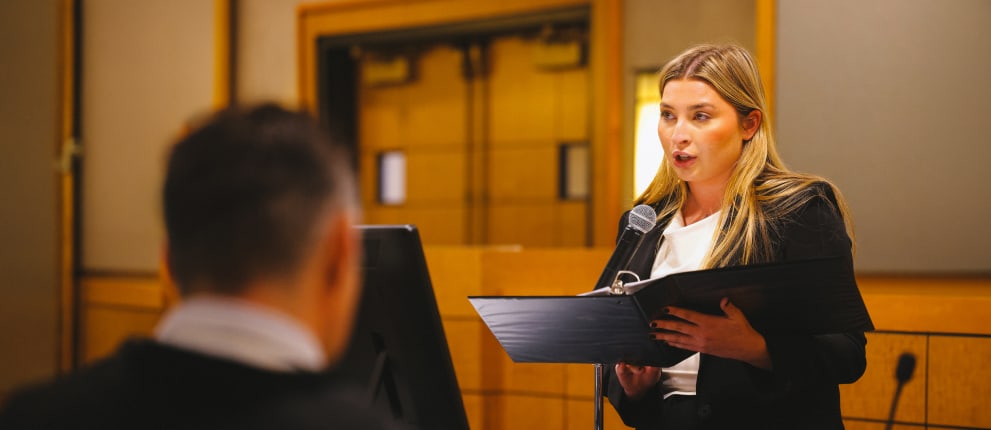 A woman in professional attire speaks into a microphone while holding a binder in a courtroom, with a seated person blurred in the foreground.