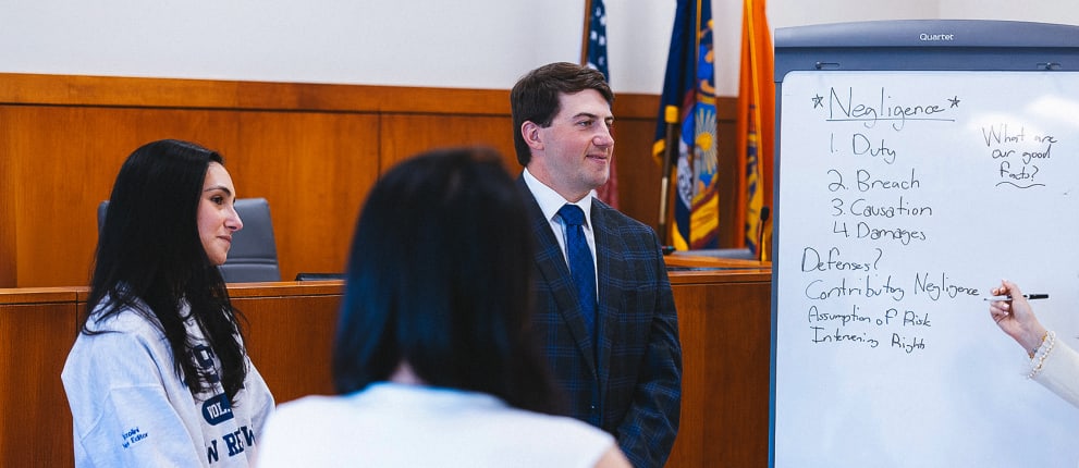 Three people stand in a courtroom; one woman writes on a whiteboard listing elements of negligence, while two others listen. Flags and wooden panels are in the background.