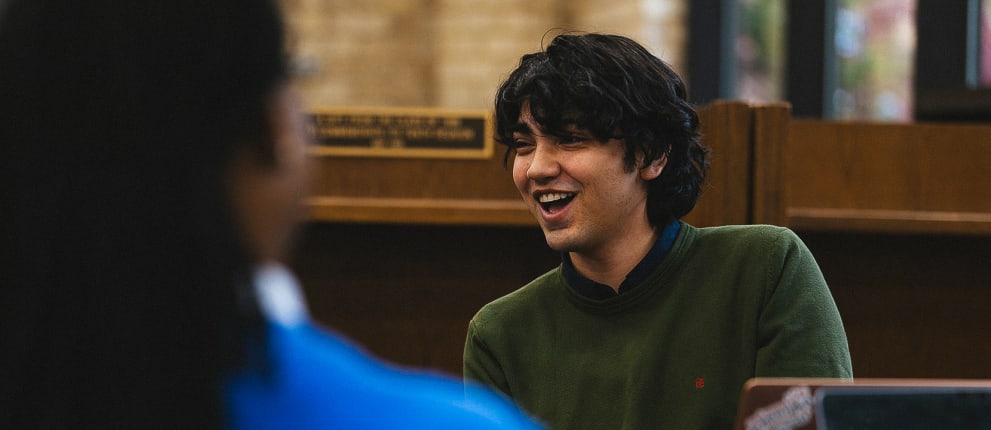 A young man with dark hair, wearing a green sweater, smiles while sitting indoors and speaking with another person who is partially visible in the foreground.