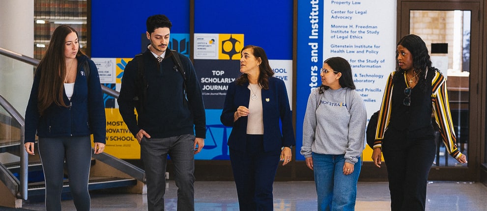Four people walk and talk together inside a building with a blue information board in the background and a staircase to the left. The group appears engaged in conversation, and all are wearing business-casual or casual attire.