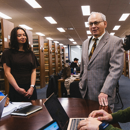 A man in a suit and a woman in a brown dress stand smiling by a table in a library, while two people work on laptops. Bookshelves and other people studying are visible in the background.