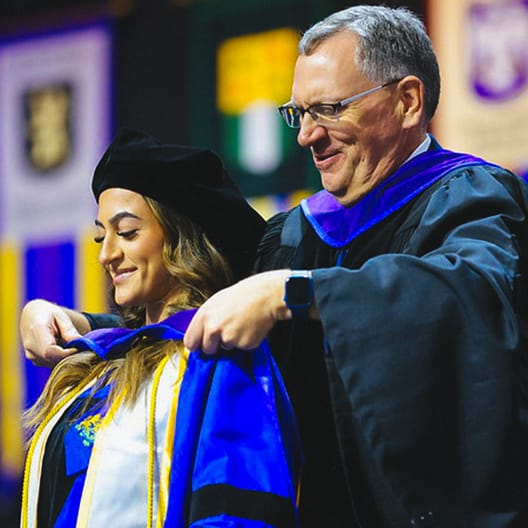 A smiling graduate in a cap and gown is being hooded by a faculty member during a graduation ceremony. Both are dressed in academic regalia, and colorful banners hang in the blurred background.