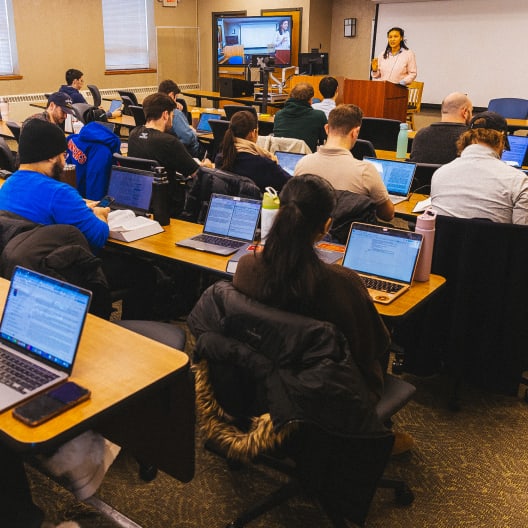 Students sit at desks with laptops open, facing a lecturer at the front of a classroom. The lecturer speaks from a podium; a screen behind shows a remote participant on a video call.