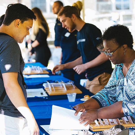 A woman sits at a table with papers, assisting a man standing across from her at an outdoor event. Other people talk and interact at nearby tables covered in blue tablecloths.
