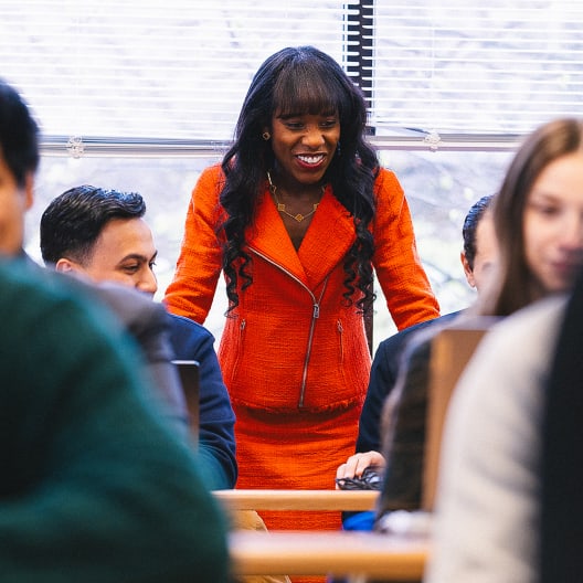 A woman in a bright orange jacket stands and smiles while engaging with people seated at desks in a classroom or office setting.
