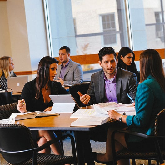 Three people in business attire sit at a table with open books and a tablet, engaged in discussion. Others work at nearby tables in a bright, modern office or classroom with large windows.