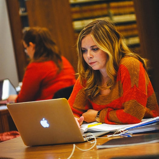 A woman with long blonde hair, wearing an orange sweater, works on a laptop at a desk with notebooks and papers. Another person is visible in the background, also using a laptop.
