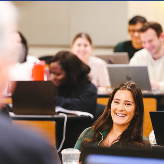 A young woman smiling in a classroom, surrounded by other students sitting at desks with laptops open. The atmosphere appears positive and engaged.