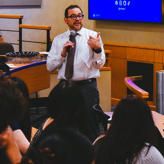A man in a white shirt and striped tie speaks into a microphone while standing in front of an audience in a lecture hall, with a large screen displaying icons behind him.