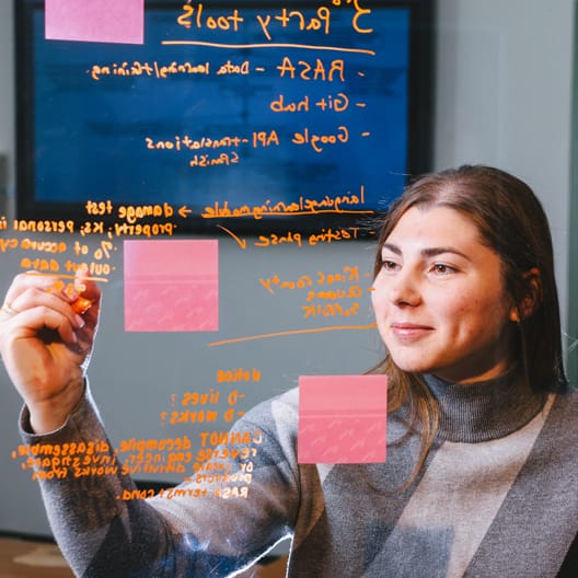 A woman writes notes with an orange marker on a glass board covered in sticky notes and text, focusing intently on her work in a modern office setting.
