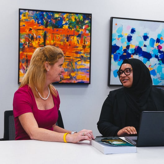 Two women sit at a table having a conversation; one wears a pink dress, the other a black hijab and glasses with a laptop. Colorful abstract paintings hang on the wall behind them.