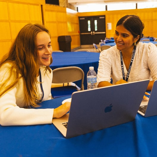 Two women sit at a table with blue tablecloths, smiling and talking while working on laptops. One has long light hair, the other has dark hair tied back. A water bottle and chairs are visible in the background.