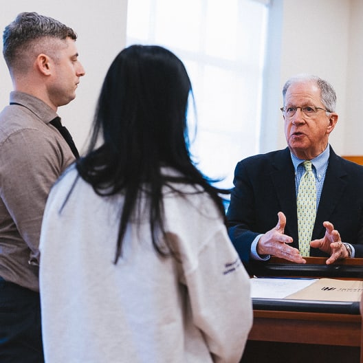 An older man in a suit speaks emphatically to a young man and woman standing across from him at a wooden desk in a brightly lit room.