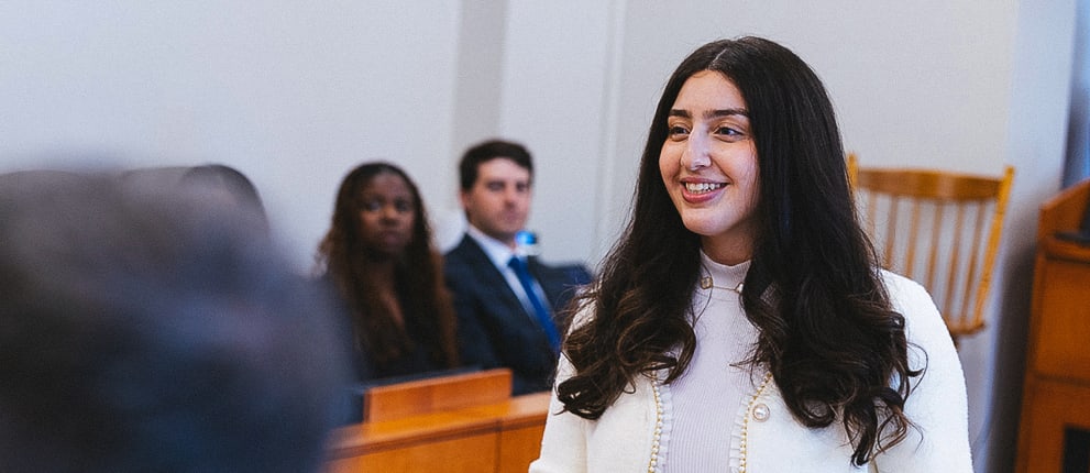 A woman with long dark hair smiles while standing and speaking in a room with seated people and wood furniture, possibly a courtroom or lecture hall.