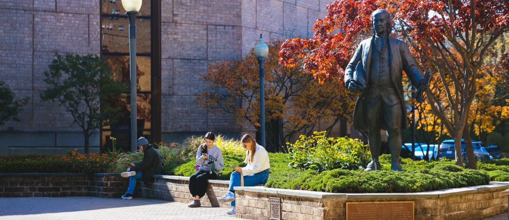 Three people sit on a low brick wall by a statue in a landscaped campus area with green shrubs and yellow flowers on a sunny day. Two are reading or using phones, and one sits quietly. Trees with autumn leaves are in the background.