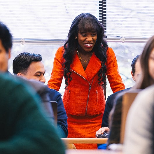 A woman in an orange suit stands smiling and engaged with people seated around her in a bright, indoor setting, possibly during a meeting or class.