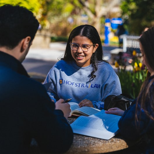 A student wearing a Hofstra Law sweatshirt smiles while studying with two others outdoors at a round table, surrounded by books and greenery.