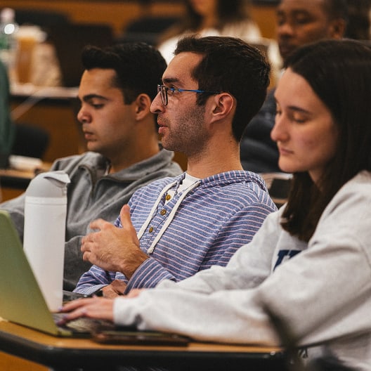 Three students sit at desks in a classroom, focused on a lecture. One student gestures while speaking, another looks at a laptop, and the third faces forward attentively. Other people are visible in the blurred background.