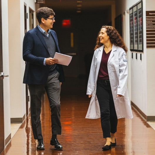 A man in a suit and a woman in a white lab coat walk together down a hallway, smiling and talking. The man holds papers, and they appear to be in a professional or academic setting.