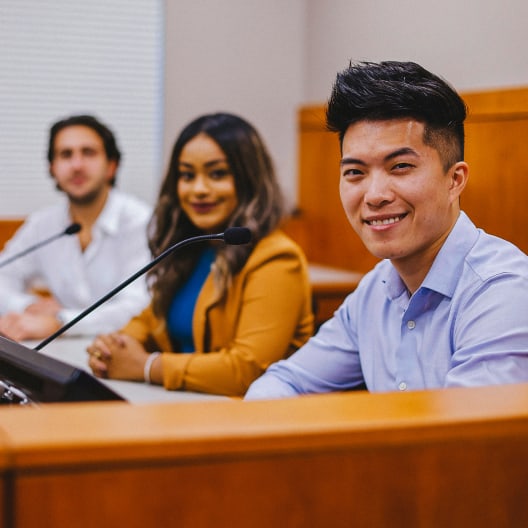 Three people sit at a conference table with microphones, facing forward. The person in the front is smiling at the camera; the others are seated behind, appearing attentive. The setting looks like a meeting or courtroom.