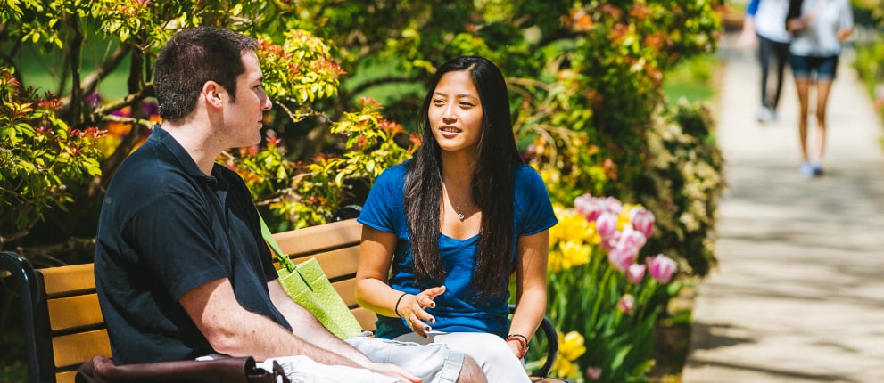 Two people sit on a bench in a garden, engaged in conversation. Colorful flowers and greenery surround them, and a path with people walking is visible in the background.