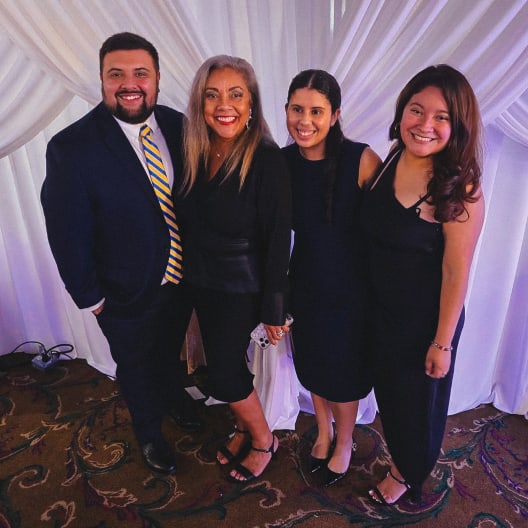 Four people dressed formally, standing close together and smiling at the camera in front of white draped curtains. The group consists of one man in a suit and three women in black dresses.