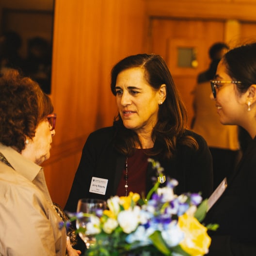 Three women are engaged in conversation at an indoor event. One woman holds a glass, and a bouquet of flowers is visible in the foreground. The background is softly lit, giving a warm atmosphere.