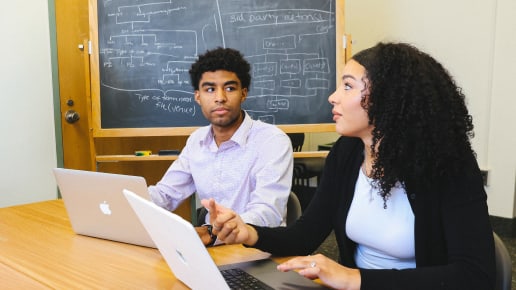 Two people sit at a table with laptops, engaged in conversation. A chalkboard with diagrams and text is in the background. The setting appears to be a classroom or study room.