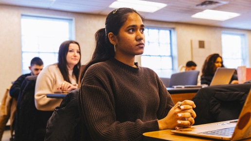 A young woman sits attentively at a desk in a classroom, with a laptop in front of her. Other students are seated nearby, also engaged in the class. Natural light comes through large windows in the background.