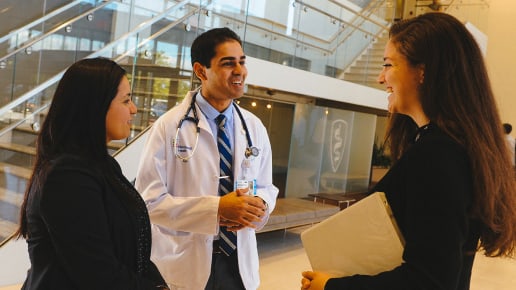 Two women and a male doctor with a stethoscope smile and talk together in a bright, modern building lobby; one woman holds a closed laptop.
