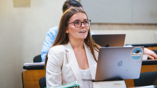 A woman wearing glasses and a white blazer sits at a desk in a classroom, looking attentively ahead. She has an open laptop in front of her and other students are visible in the background.