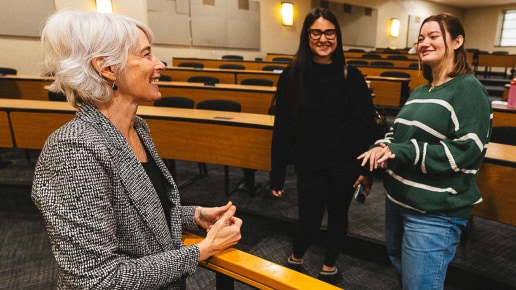 A woman with short gray hair talks and smiles with two students in an empty classroom. The students appear engaged and happy, standing near the front row of desks.