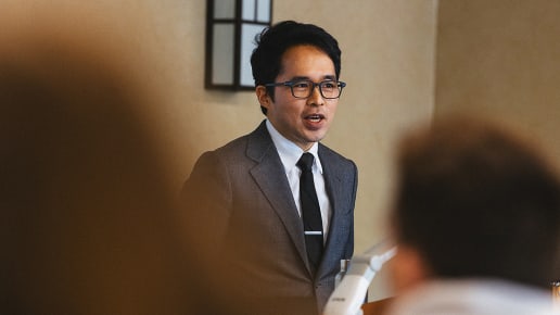 A man in a suit and tie, wearing glasses, speaks at a podium in a room with beige walls. People in the foreground listen to him attentively.