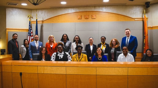 A group of fifteen professionally dressed people, both men and women, pose together in a courtroom behind a wooden bench. The American and state flags are visible in the background.