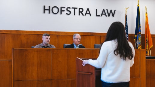 A woman stands at a podium speaking to two men seated behind a judge’s bench in a courtroom setting with the words Hofstra Law on the wall and flags in the background.