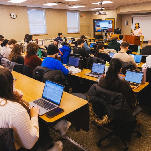 A classroom with students seated at desks using laptops, facing a lecturer at the front near a podium and a screen displaying a presentation. The room has beige walls, windows, and fluorescent lighting.