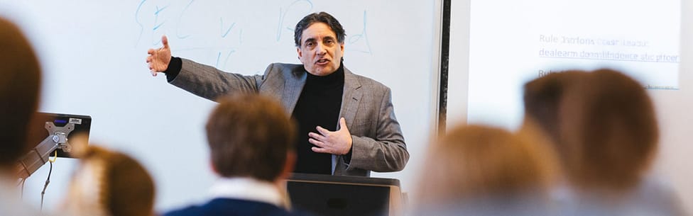 A man in a gray blazer gestures while speaking in front of a classroom, addressing students seated facing him. A whiteboard and a projected slide are visible behind him.