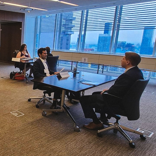 A man and a woman in business attire stand smiling in front of large windows with a city skyline in the background.