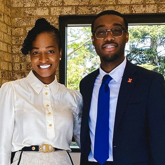 Two people stand indoors smiling at the camera. The woman is wearing a white blouse and a patterned belt, and the man is in a dark suit with a blue tie and glasses. There is a window and brick wall behind them.