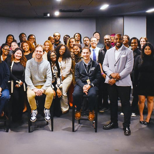 A large, diverse group of people pose together indoors, with some sitting in front and others standing behind, all smiling at the camera in a well-lit room.