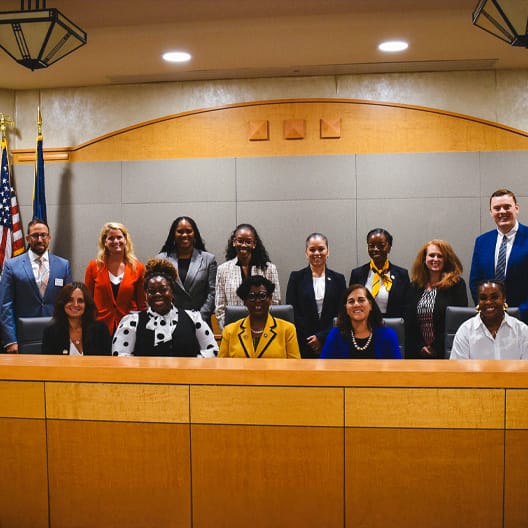 A group of 14 professionally dressed people pose and smile together in a wood-paneled courtroom or government meeting room, with flags and official decor visible in the background.