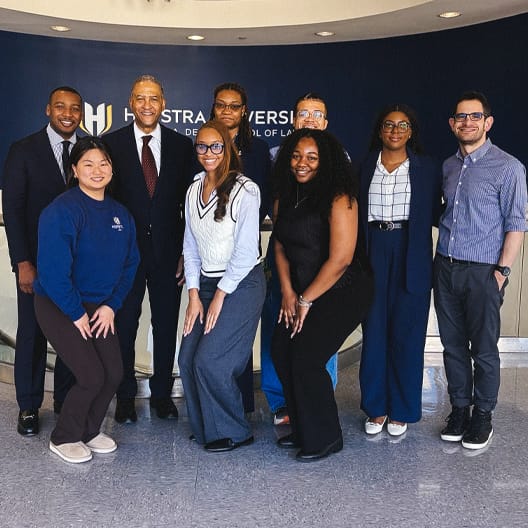A group of nine people, dressed in business and casual attire, pose and smile together indoors in front of a wall with the Hofstra University School of Law logo.