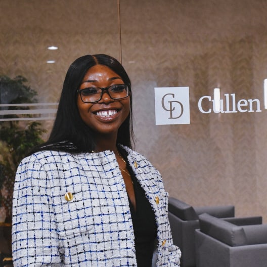 A woman with long dark hair and glasses smiles while standing in an office lobby with Cullen D on the glass wall behind her. She wears a light-colored textured jacket over a black top.