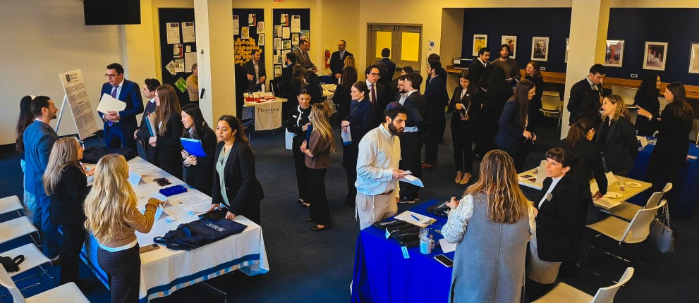 A busy career fair with people dressed in business attire networking at tables, exchanging documents, and speaking with recruiters in a well-lit room with posters and displays on the walls.