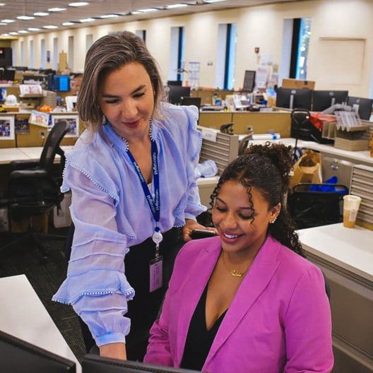 Two women in business attire work together at a computer in a modern, open-plan office. One is standing and pointing at the screen, while the other is seated and smiling, both appear engaged and collaborative.