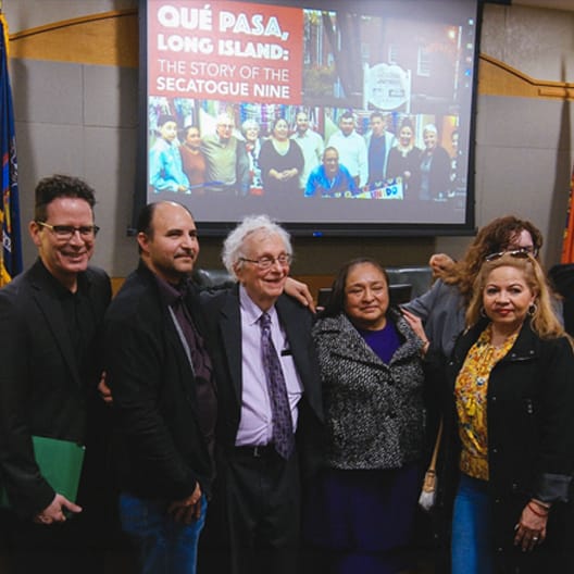A group of people stand together smiling in front of a projector screen displaying QUÉ PASA, LONG ISLAND: THE STORY OF THE SECATOGUE NINE with photos and text. They appear to be at an indoor event or presentation.