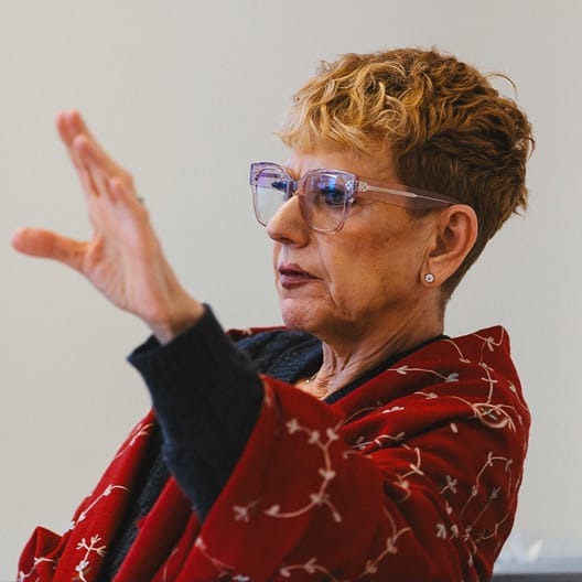 Professor Barbara Barron with short curly hair and clear glasses gestures with her hand raised. She wears a red jacket with a floral pattern and looks focused, speaking or explaining something. The background is plain and light-colored.