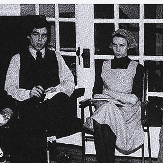 A black and white photo shows a man in a vest and tie sitting next to a woman in a long-sleeve dress with an apron and a headscarf. Both are seated indoors in front of glass doors, facing the camera.